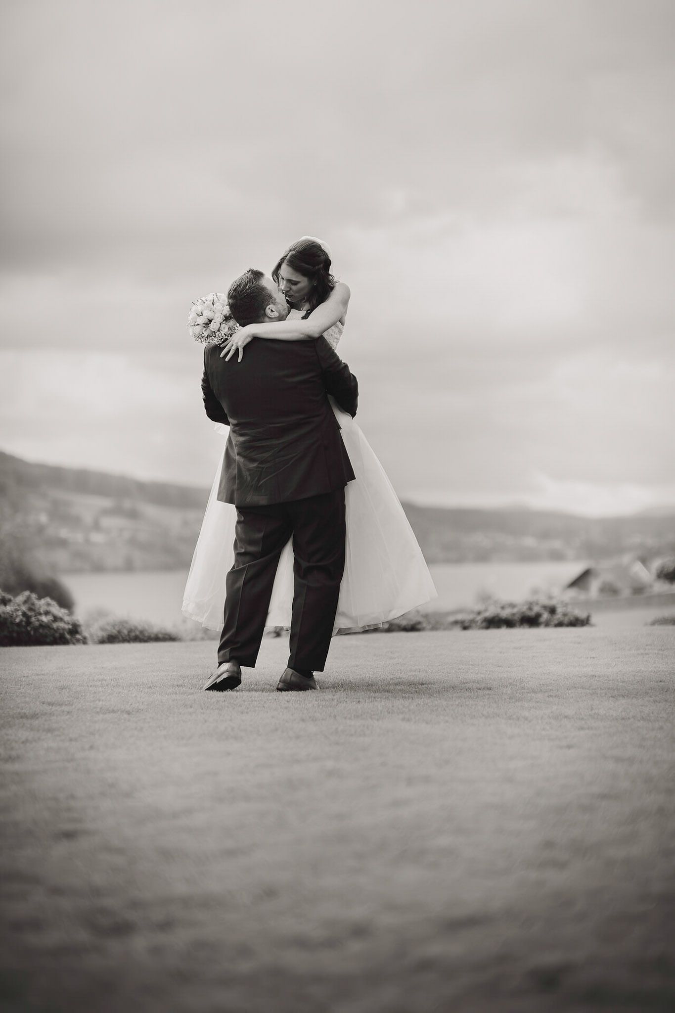A romantic wedding couple embraces on a hilltop overlooking a scenic coastal landscape.