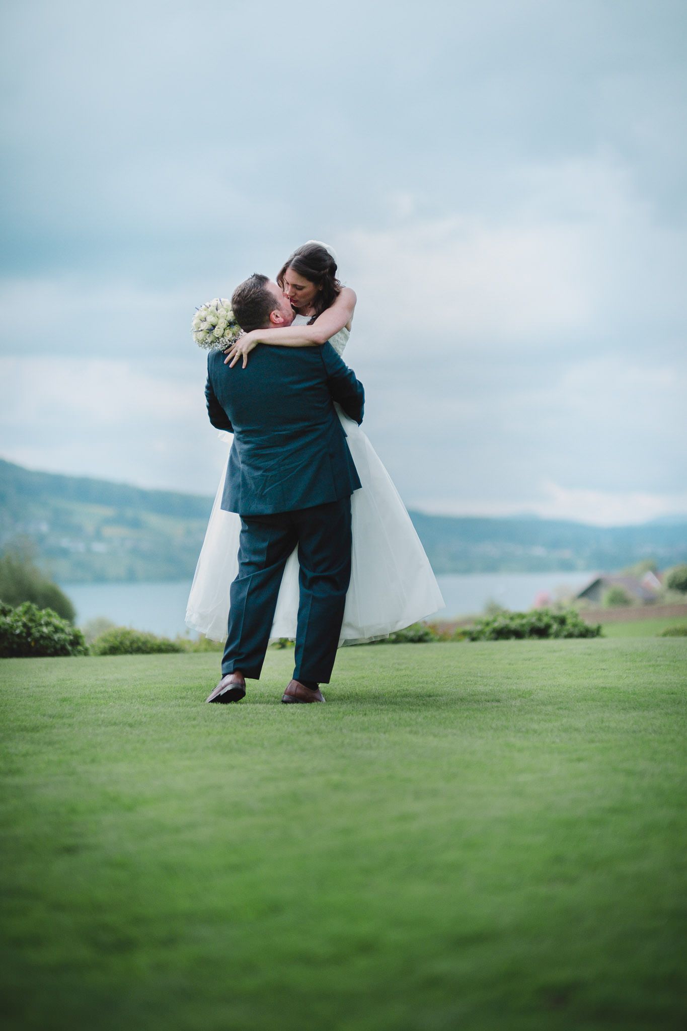 Newlyweds share an intimate moment on a grassy hillside with ocean views in the background.