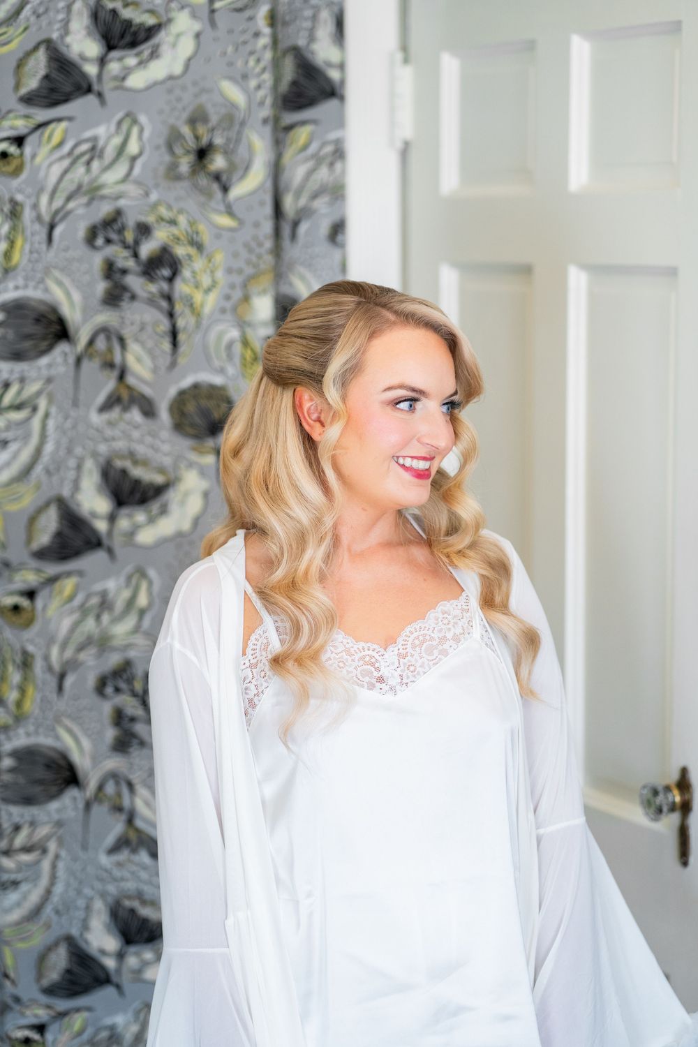 A person in a white lace top and blonde hair smiles while looking to the side against a gray patterned wallpaper background.