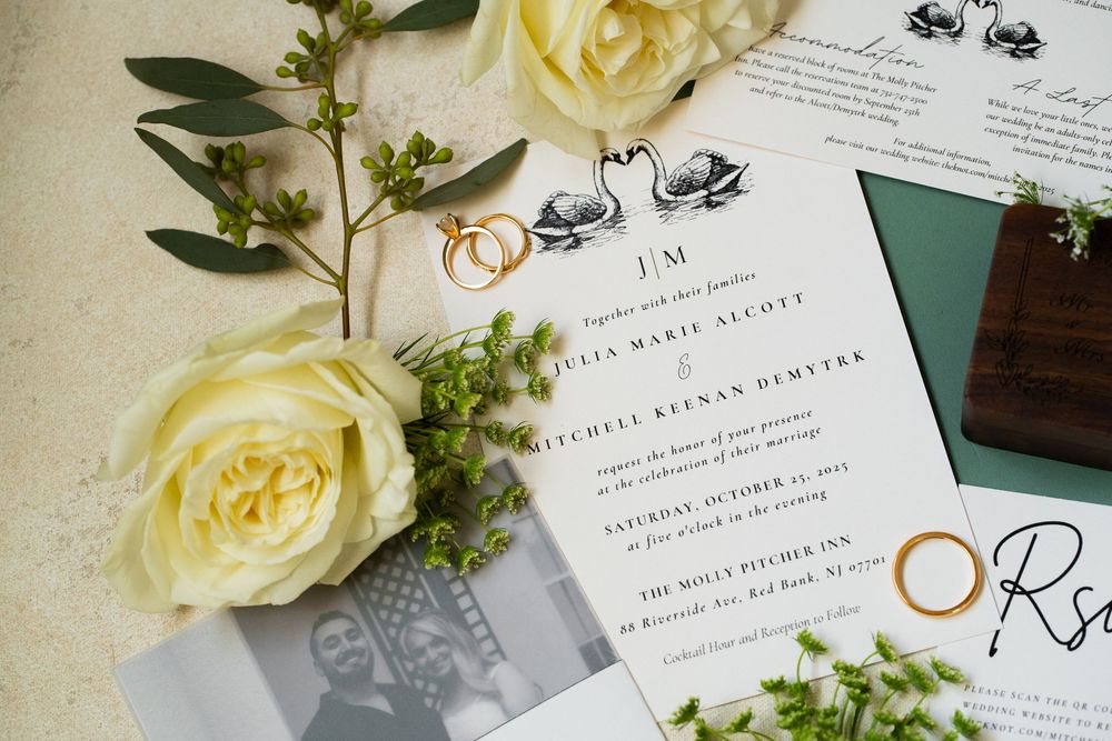 Wedding invitation with white roses, greenery, and gold wedding rings displayed on a light colored surface.
