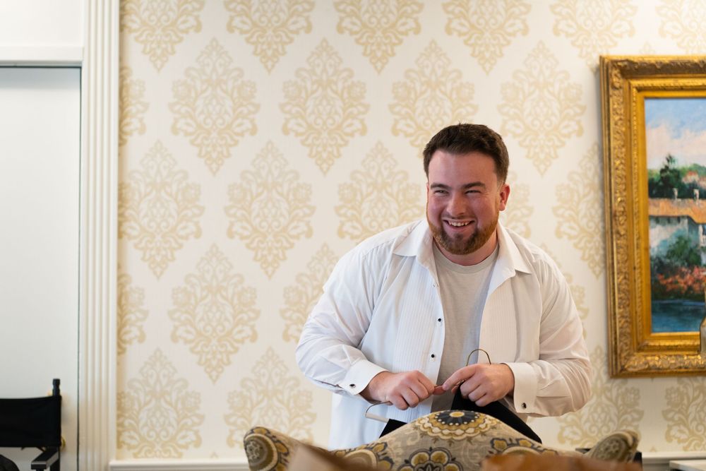 A person in a white shirt laughs while sitting at a desk in a room with ornate wallpaper and framed artwork.