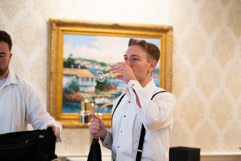 A person in formal attire adjusts their bowtie while getting ready in an elegant room with framed artwork.