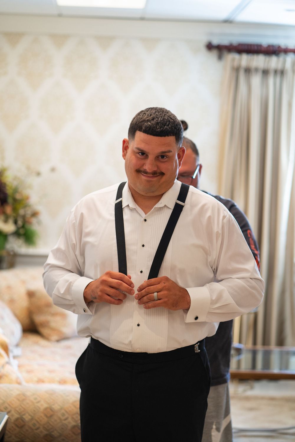 Man in white dress shirt and black suspenders smiling while getting ready for formal event.