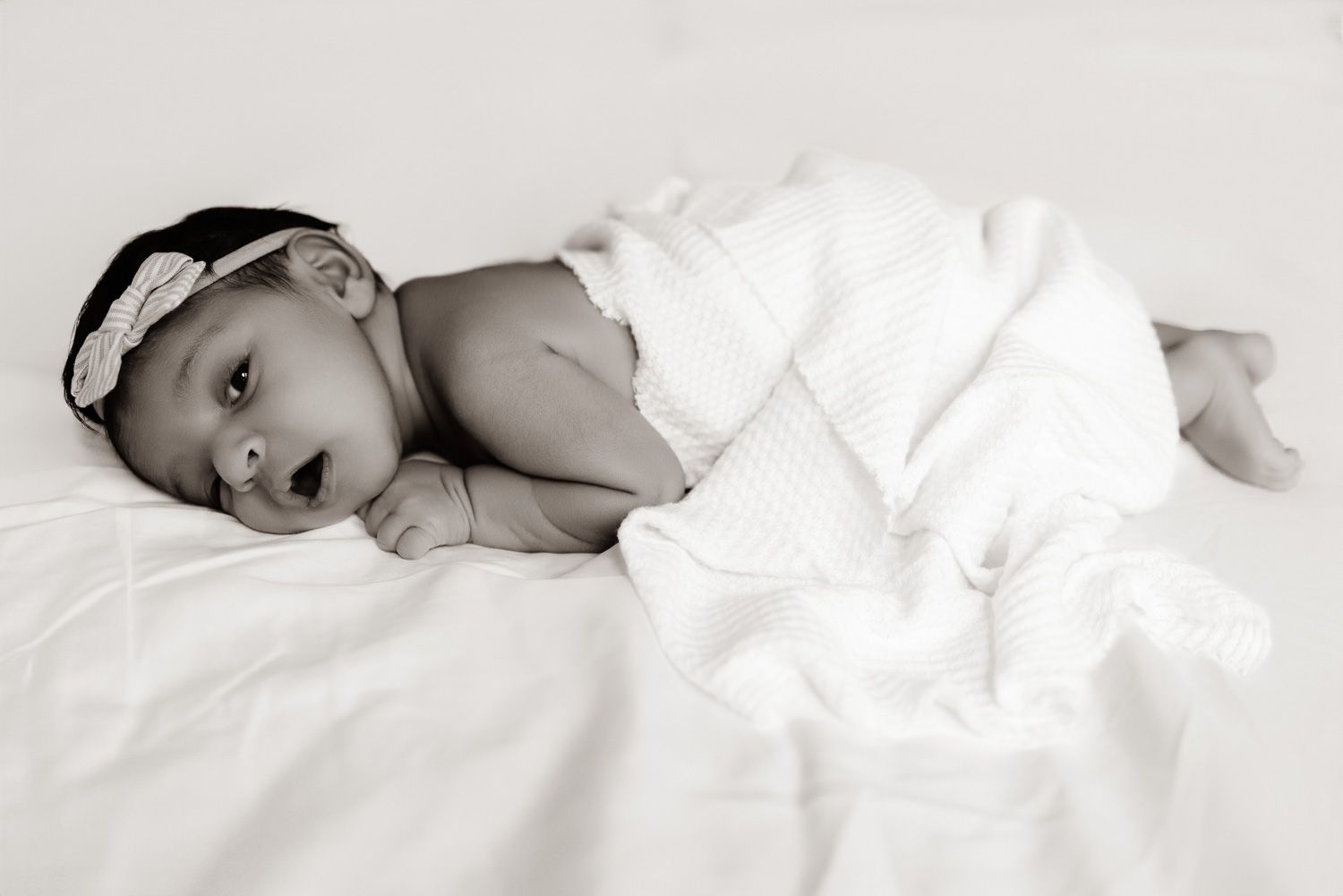 Black and white image of a newborn laying on a bed wearing a bow and swaddle