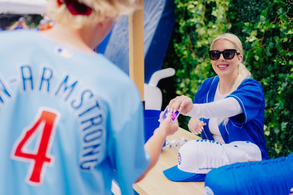 Blonde woman in blue baseball jersey smiles while signing autographs at a sports fan event outdoors.