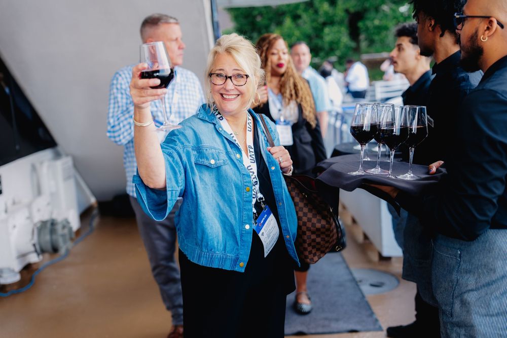 A smiling blonde woman in a denim jacket raises a glass of red wine at an outdoor networking event.