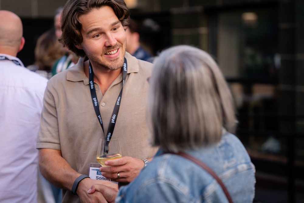 Two professionals networking at an outdoor event, man in beige polo with lanyard smiling in conversation.