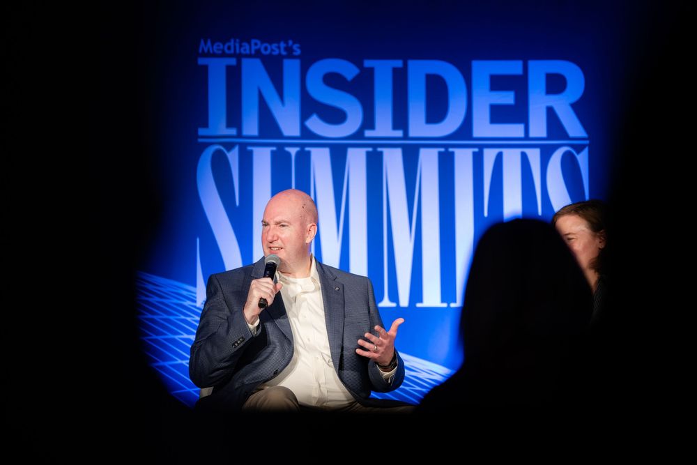 Speaker presenting at MediaPost Insider Summits conference on a blue illuminated stage with microphone.