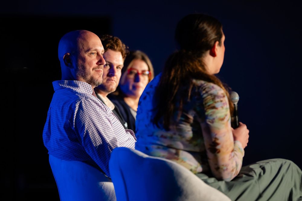 Three people seated in dim blue lighting, attentively watching an event or presentation in a darkened venue.