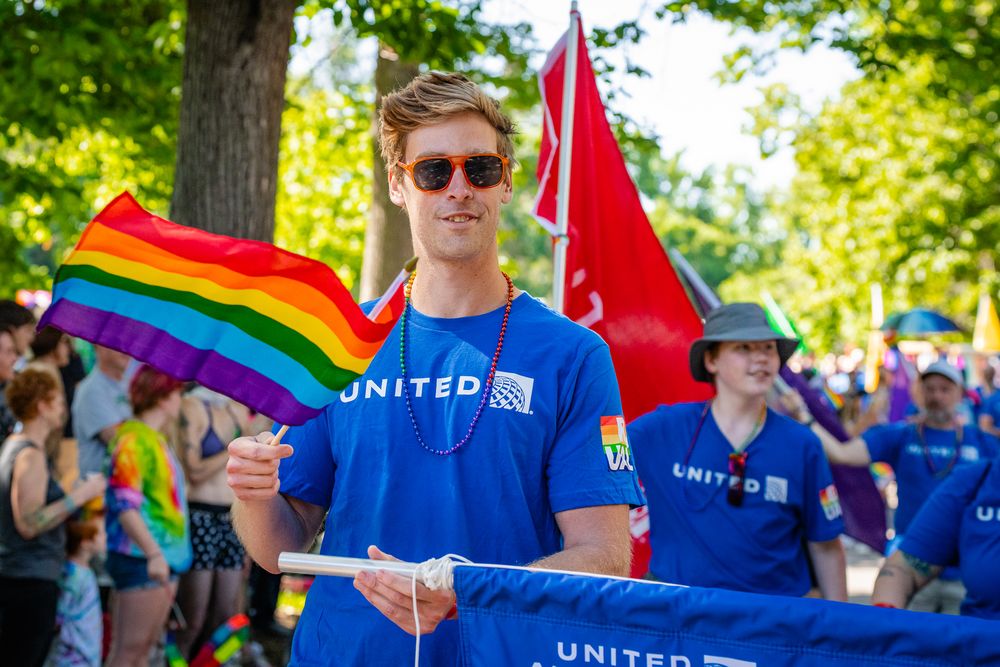 People in blue shirts at an outdoor event with rainbow pride flags and banners.