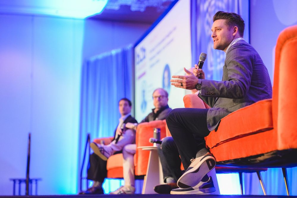 Three panelists speak on stage at a conference, seated in orange chairs under blue lighting.