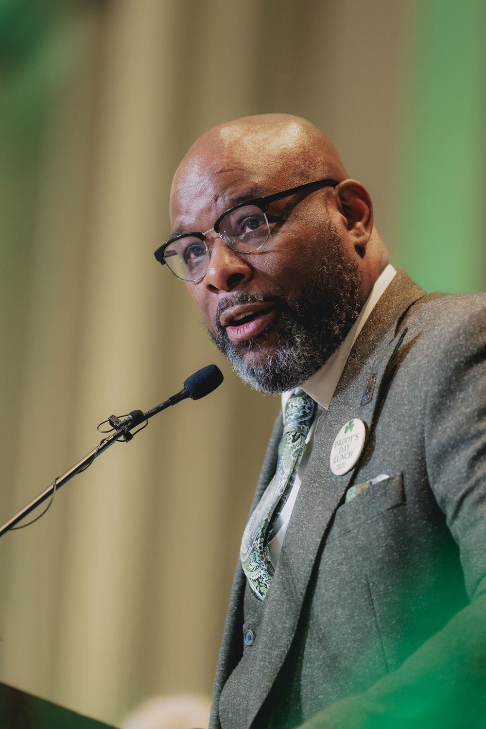 A bald man with glasses and a beard speaks at a podium microphone, wearing a gray suit with a green tie.