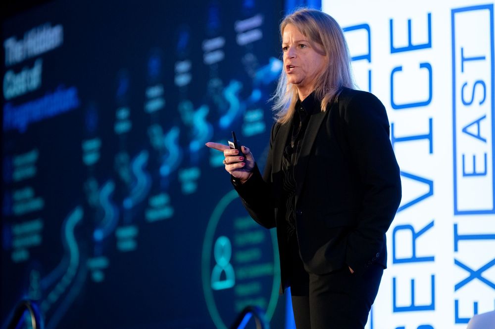 Professional woman speaker presenting at a corporate conference event with blue digital backdrop signage.