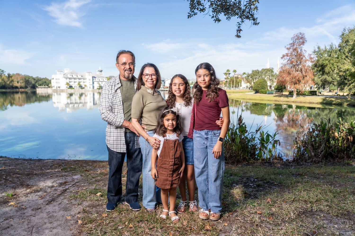 A group of people pose together outdoors near a lake during autumn with trees and buildings in the background.