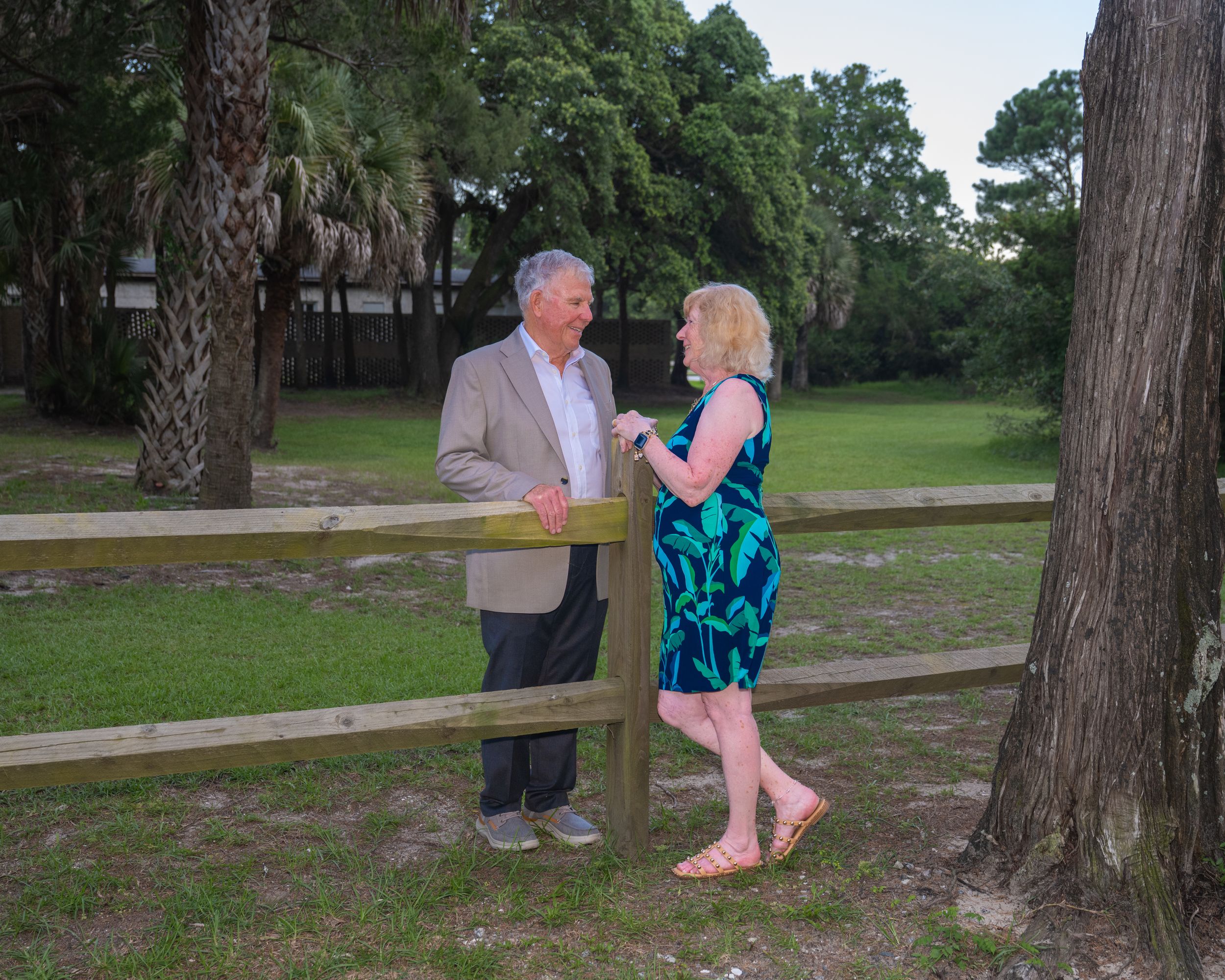 Two seniors share a sweet moment while standing beside a wooden fence in a serene park setting with tall pine trees.