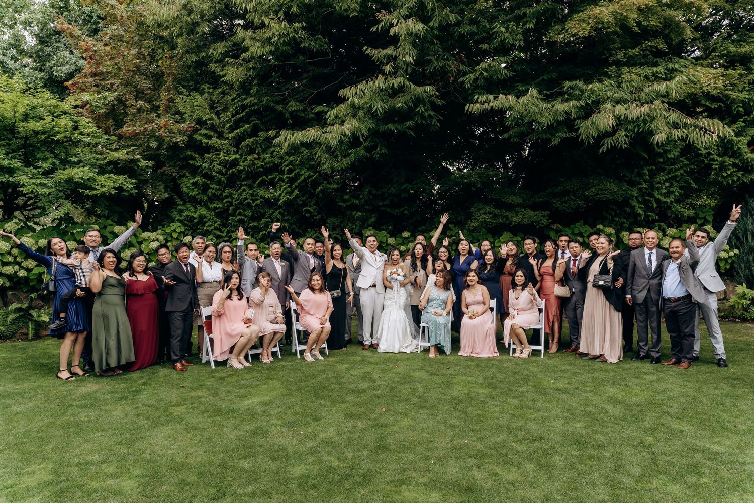 Wedding guests gathered on a green lawn for a panoramic group photo during an outdoor summer celebration.