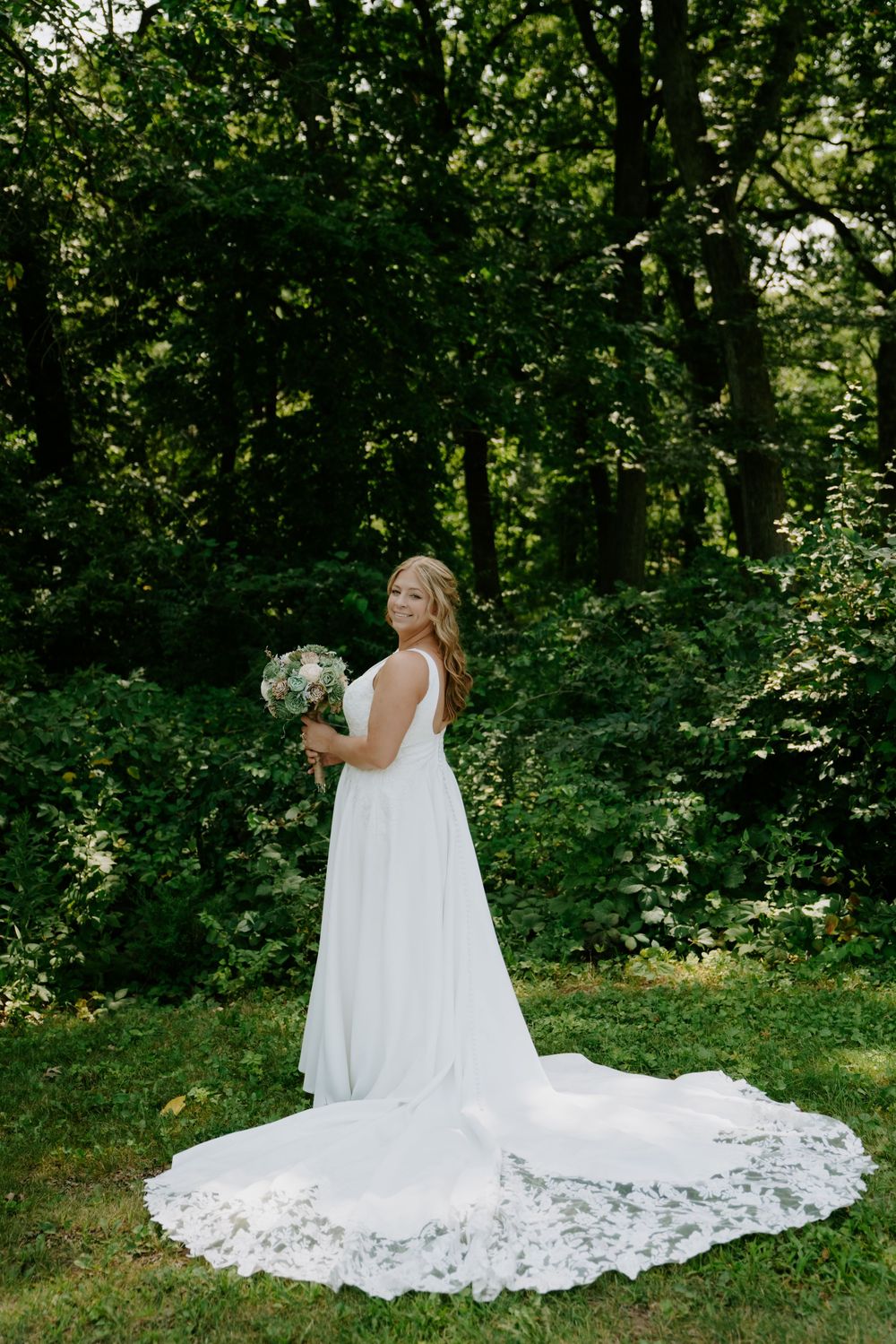 Bride in flowing white lace dress with long train poses in a lush green garden setting.