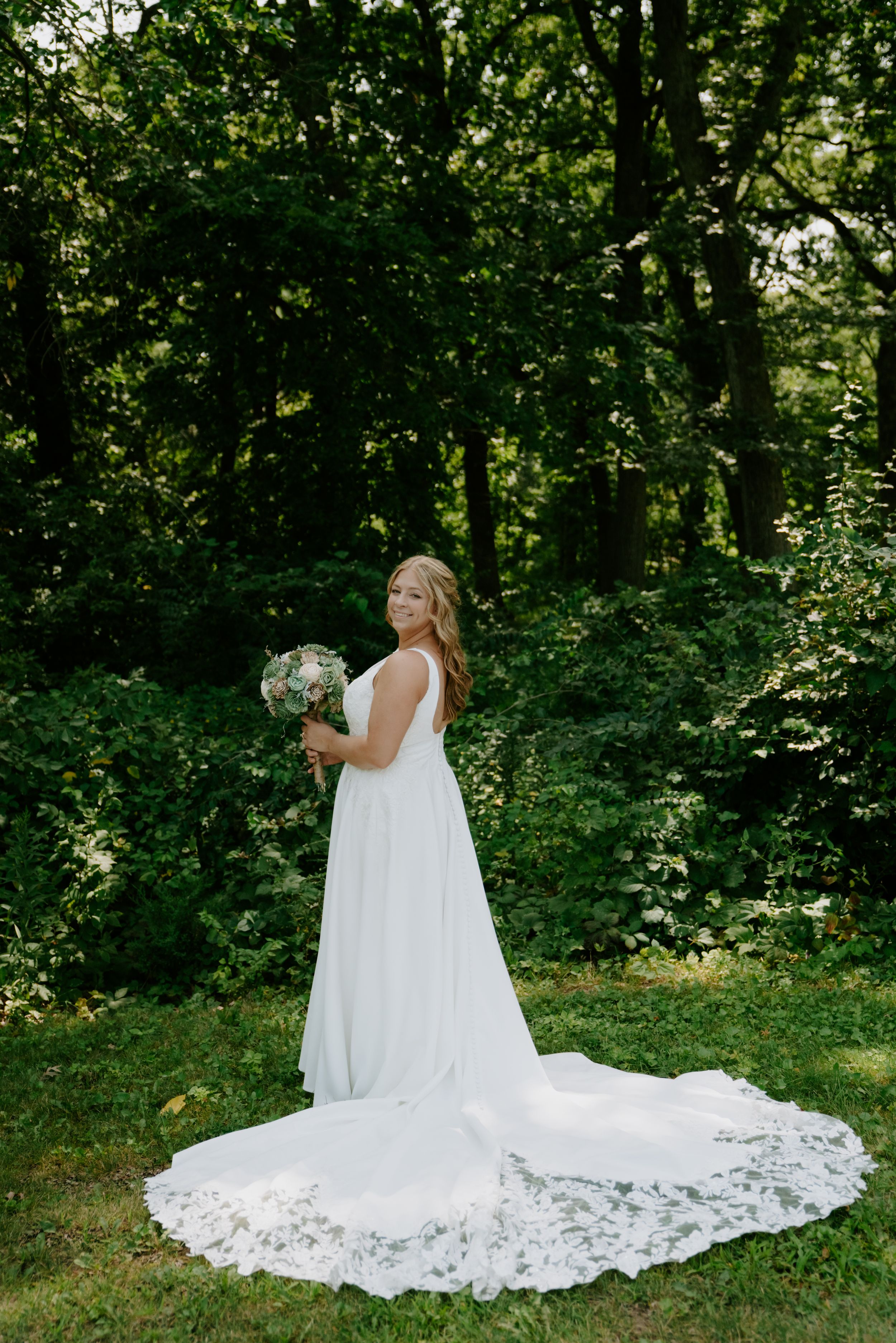 Bride in flowing white lace dress with long train poses in a lush green garden setting.