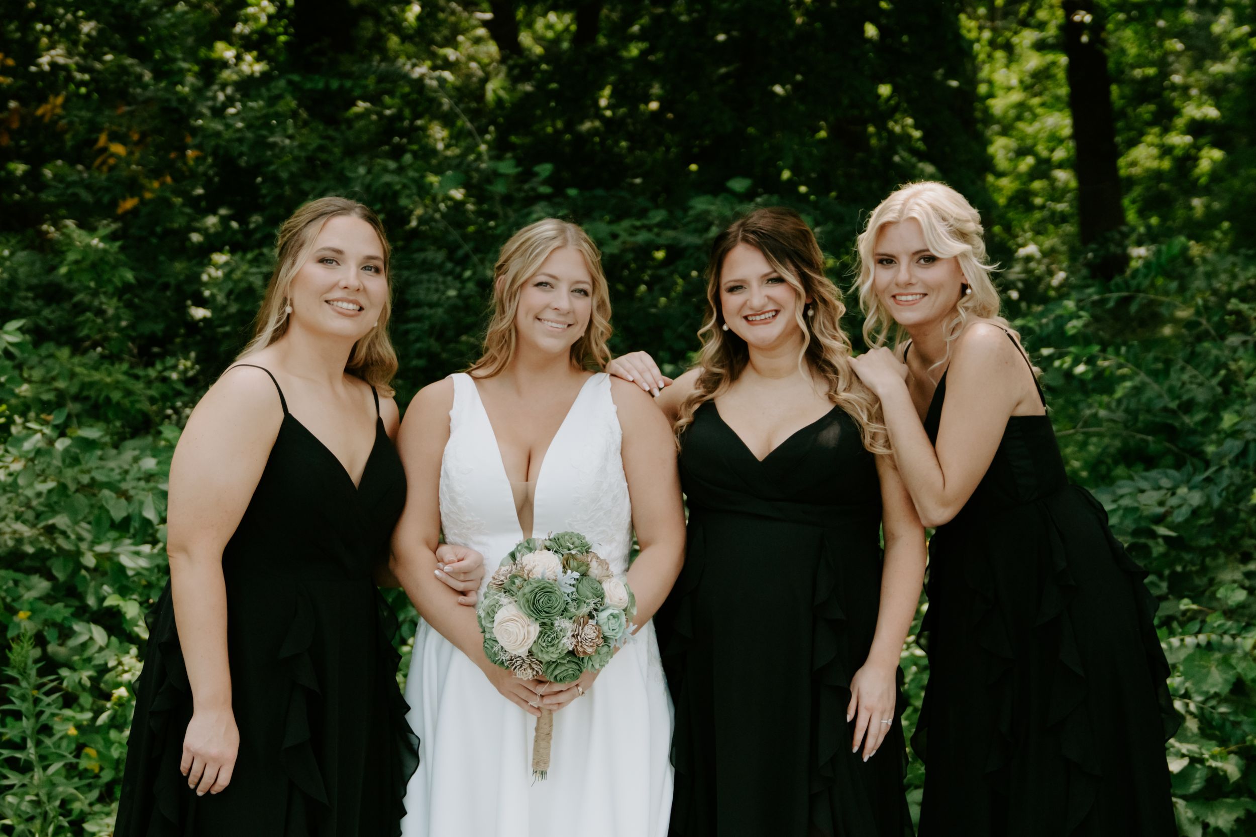 Bridal party in black dresses and white gowns posing together in a forest setting.