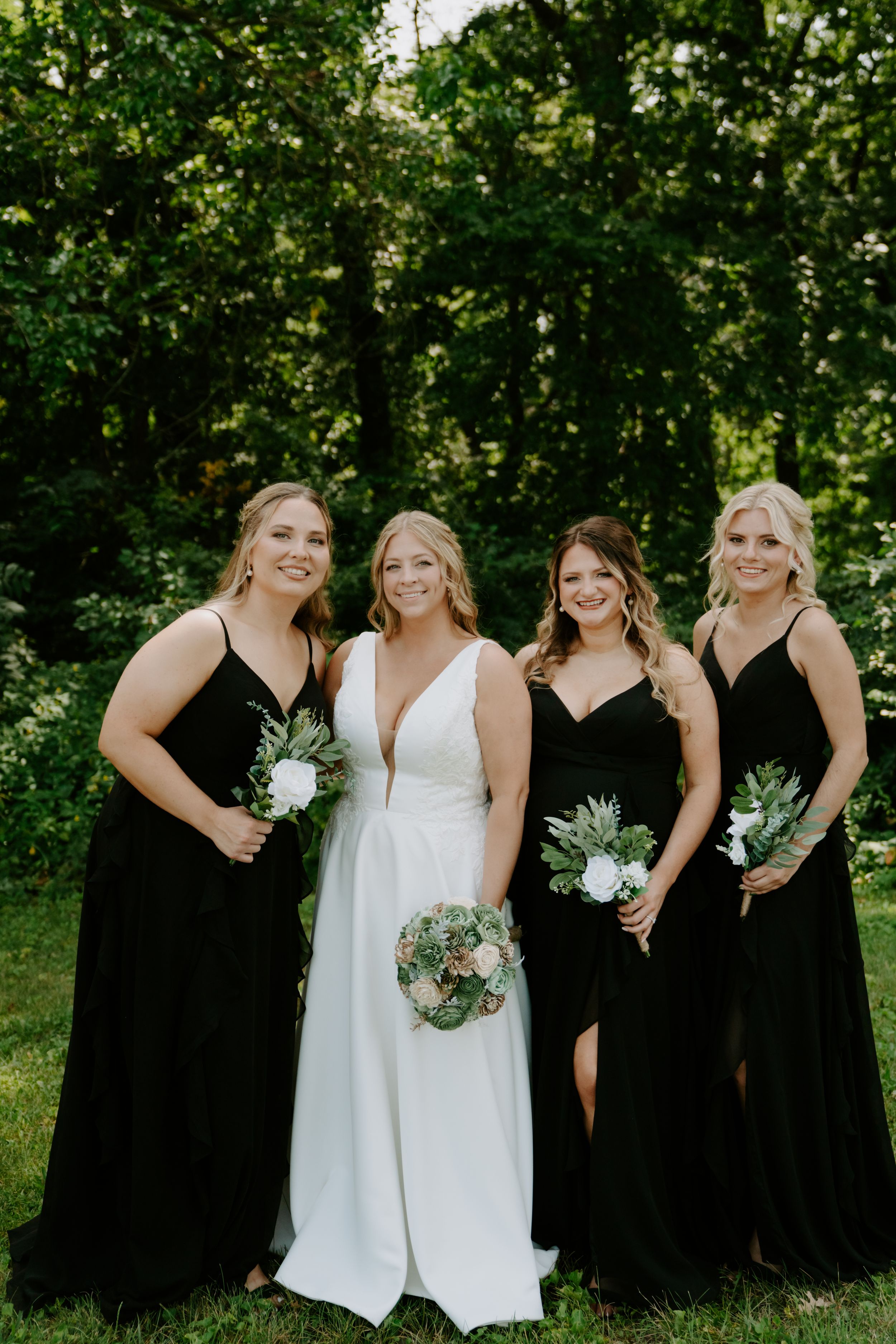 Bridal party poses outdoors with bridesmaids in black dresses and bride in white holding green and white bouquets.