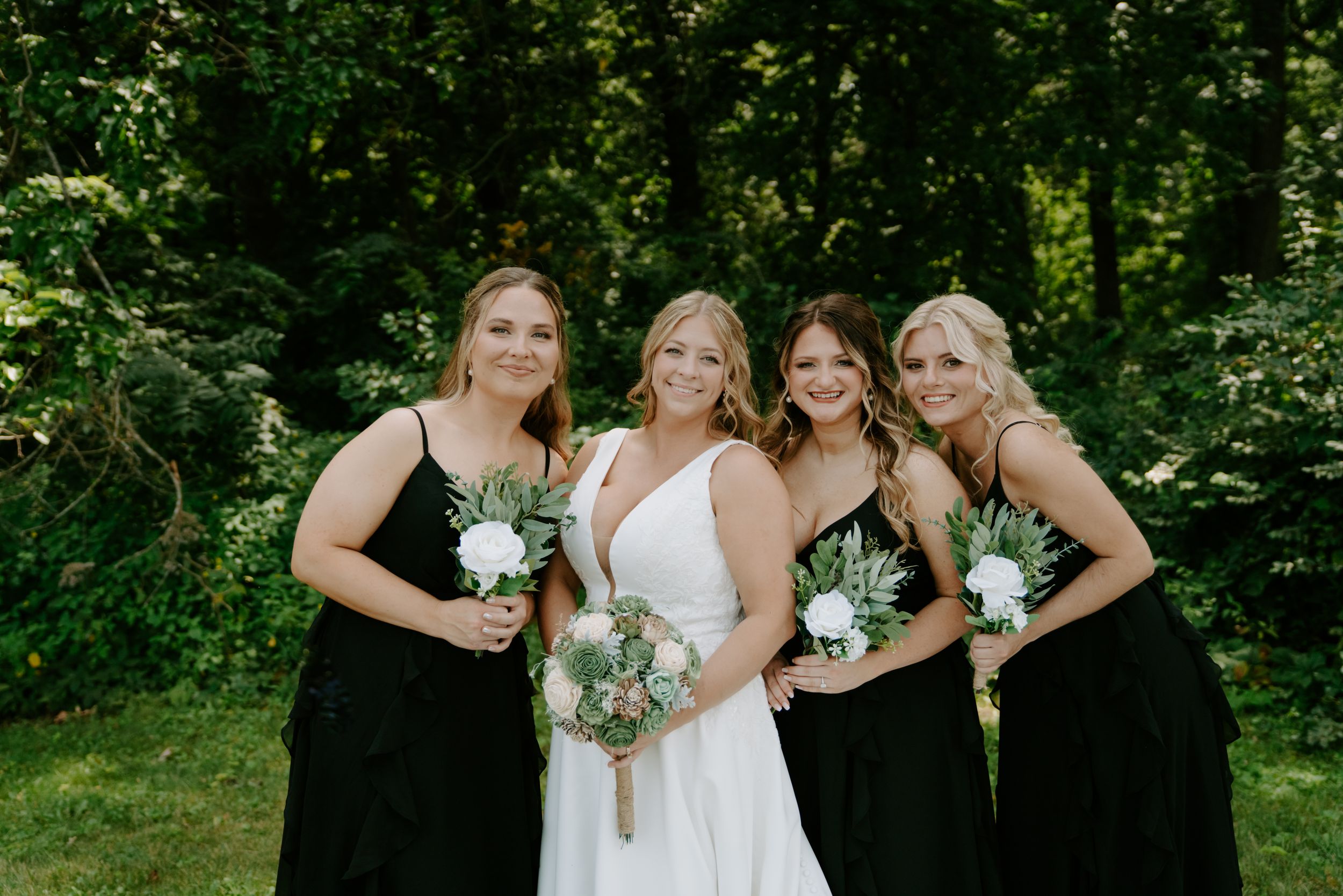 Wedding party smiles together on grass with bridesmaids in black gowns and bride in white dress.