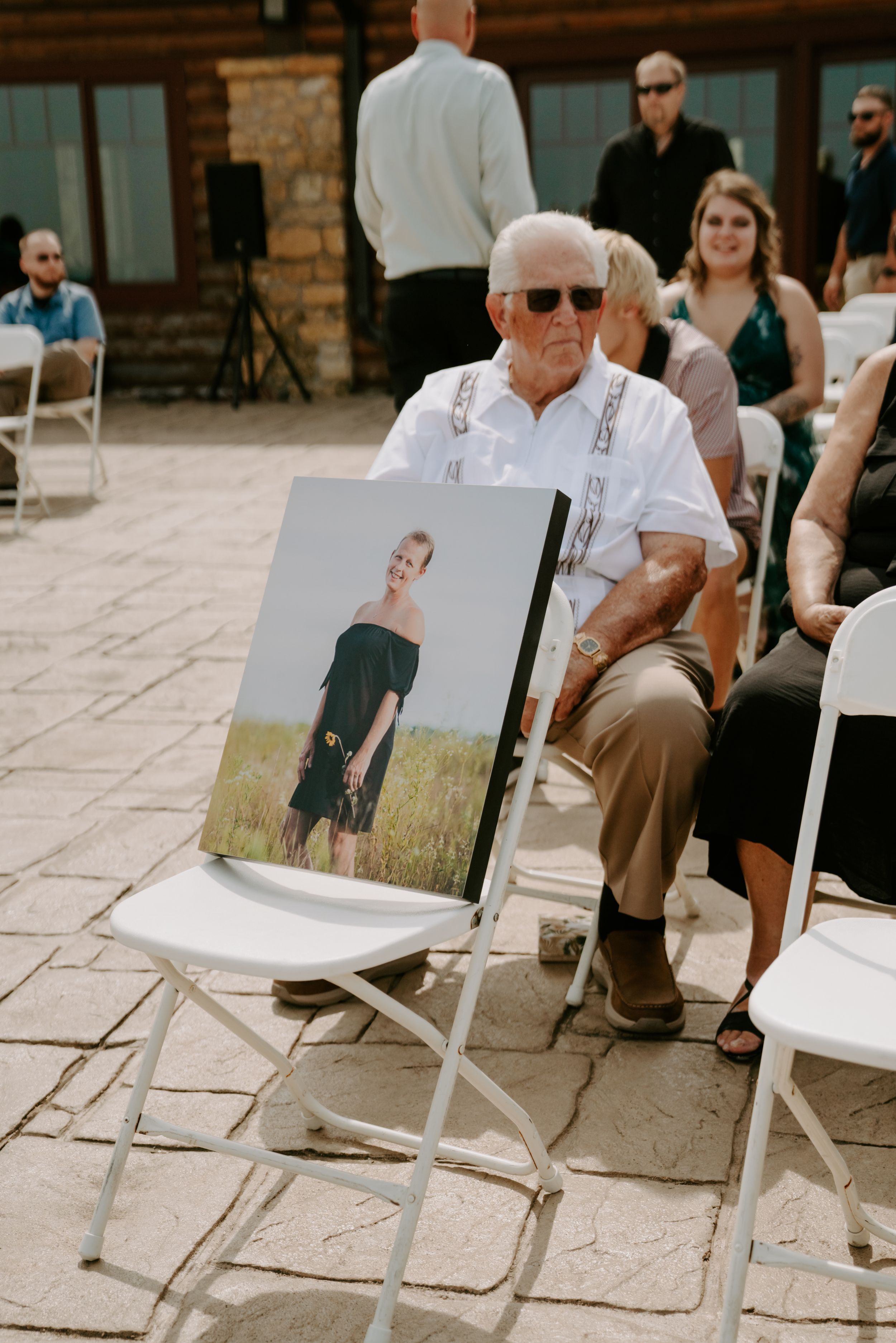 Guest in white shirt and suspenders sits next to a portrait photo display at outdoor wedding ceremony.