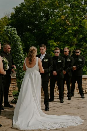 Wedding party in black attire standing on a stone terrace with trailing white wedding dress in view.