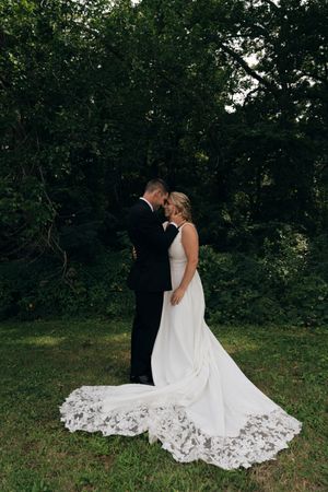 A wedding couple embracing outdoors with the bride's lace dress trailing on the grass.