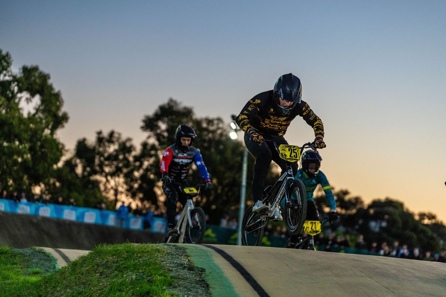 BMX riders perform tricks on a dirt track at sunset with silhouetted trees in the background.