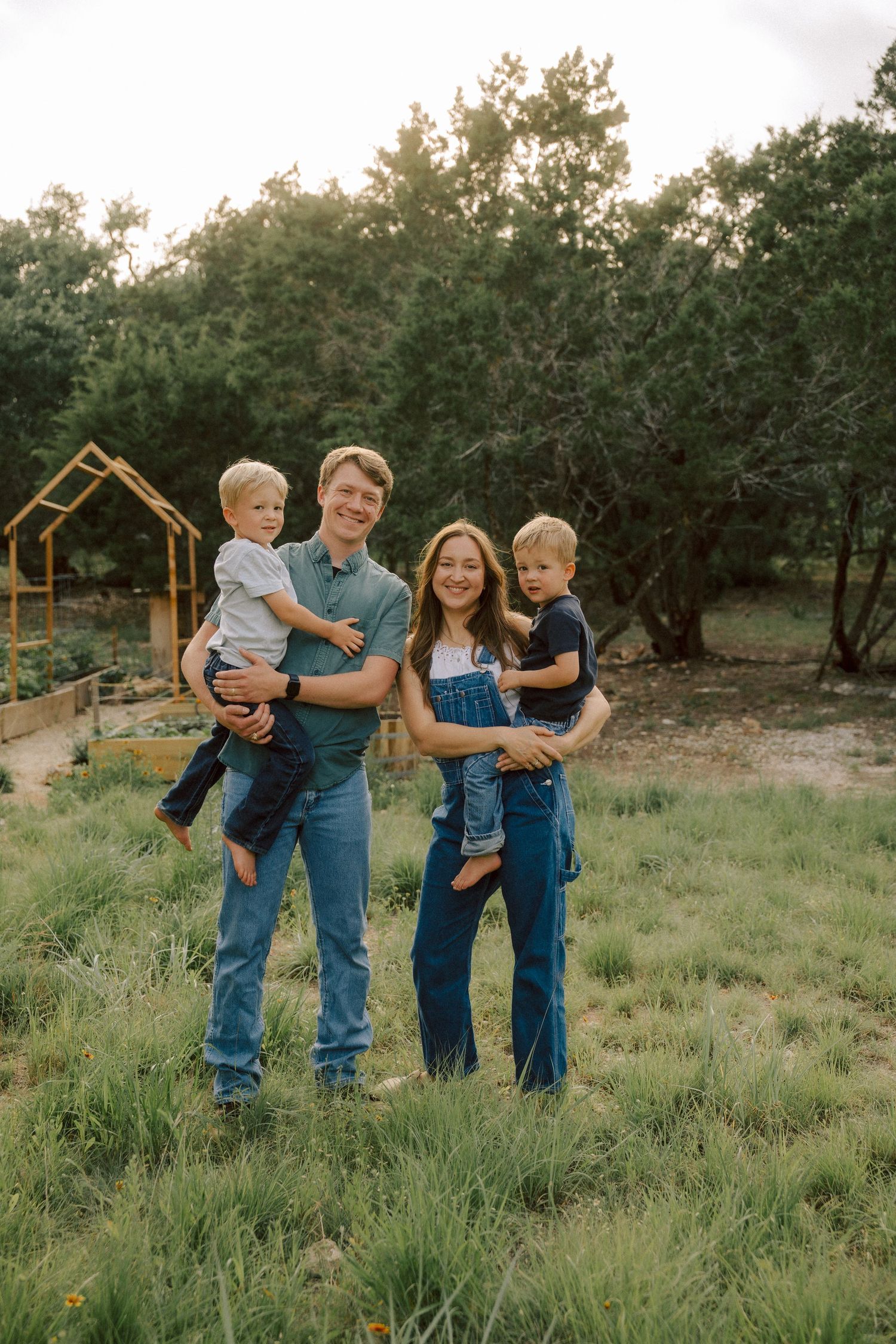 A family of four stands together in a grassy field at sunset with a rustic wooden structure visible in the background.