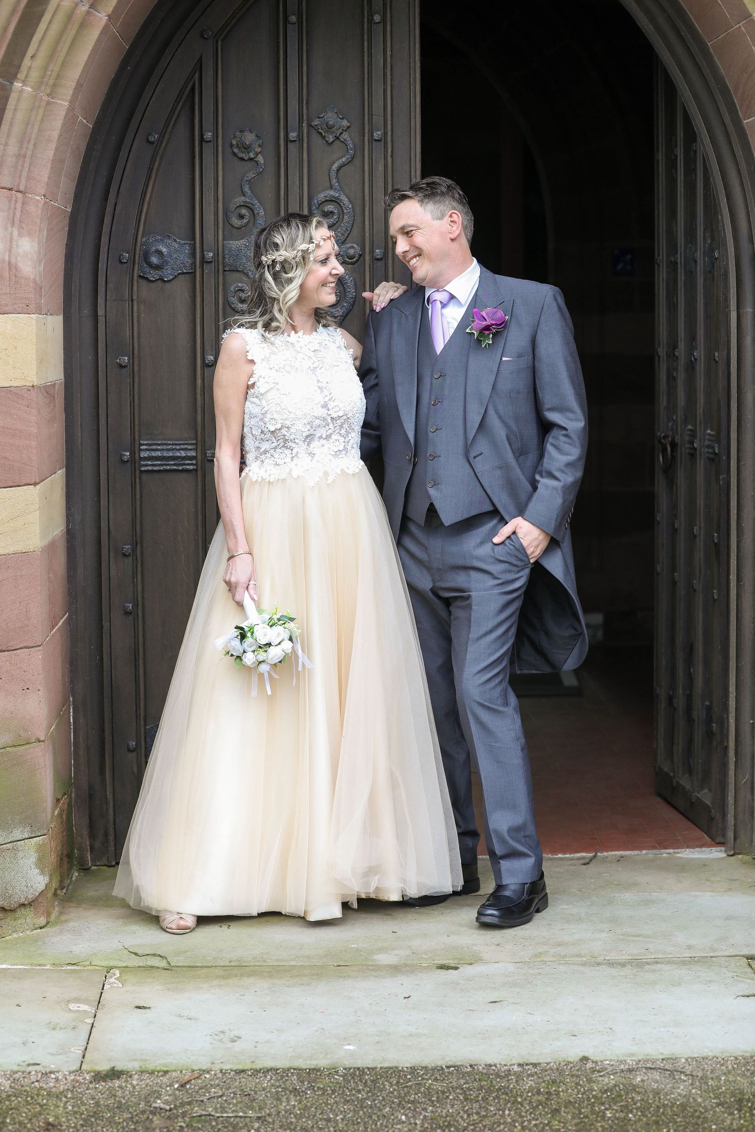 Elegant bride and groom standing in gothic church doorway wearing champagne and grey formal attire.