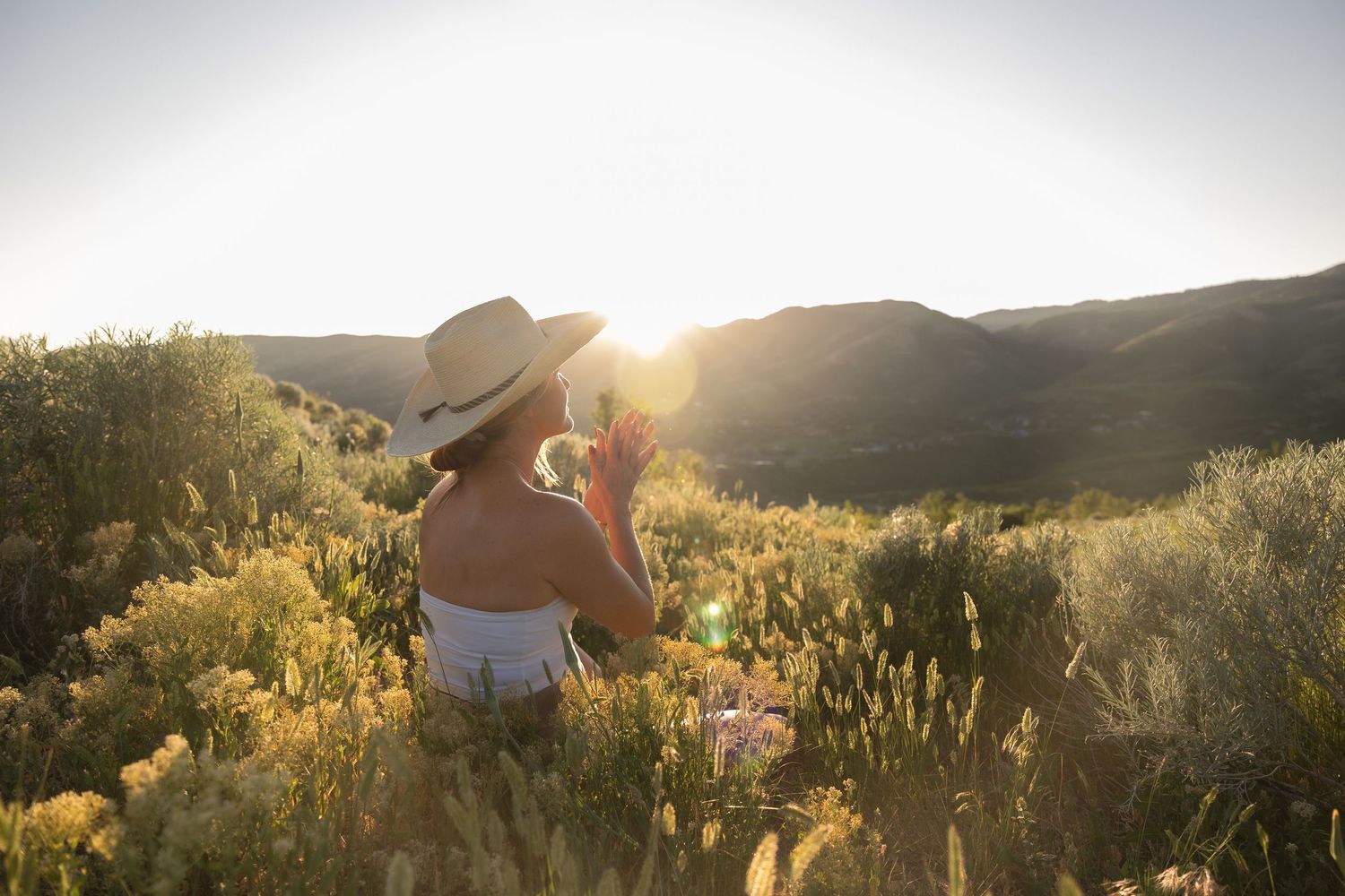 A woman meditates in golden lighting in grass with mountains in the background.