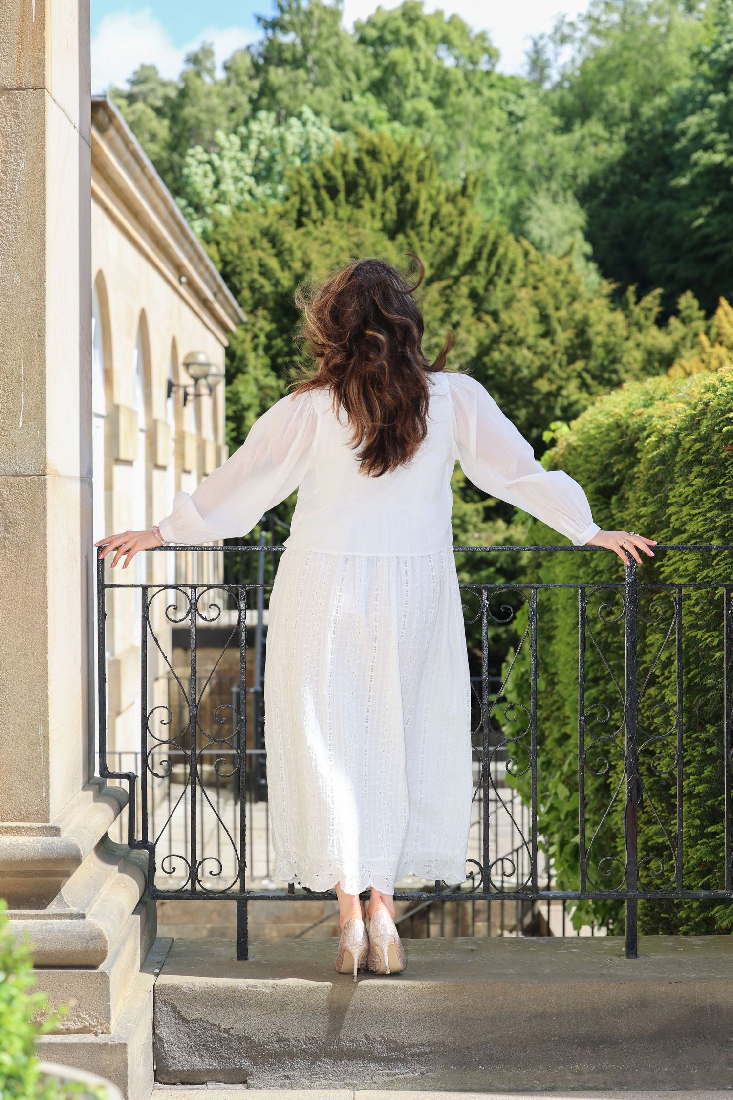 Person in flowing white long-sleeved dress stands on balcony railing with greenery and architecture in background.