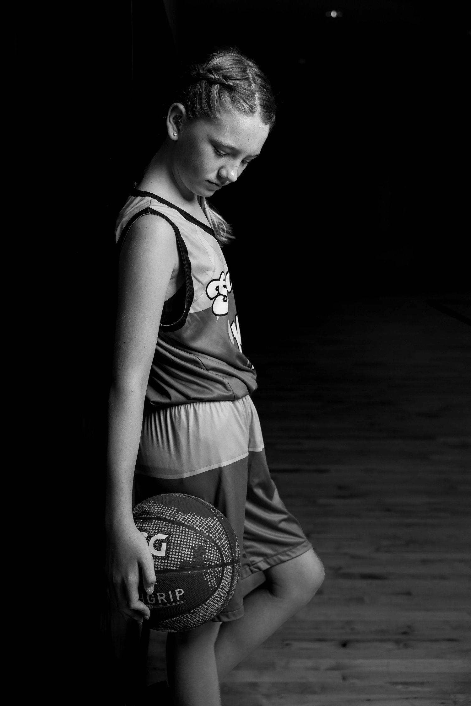 Black and white portrait of a young athlete in basketball gear holding a ball while looking down on wooden court.