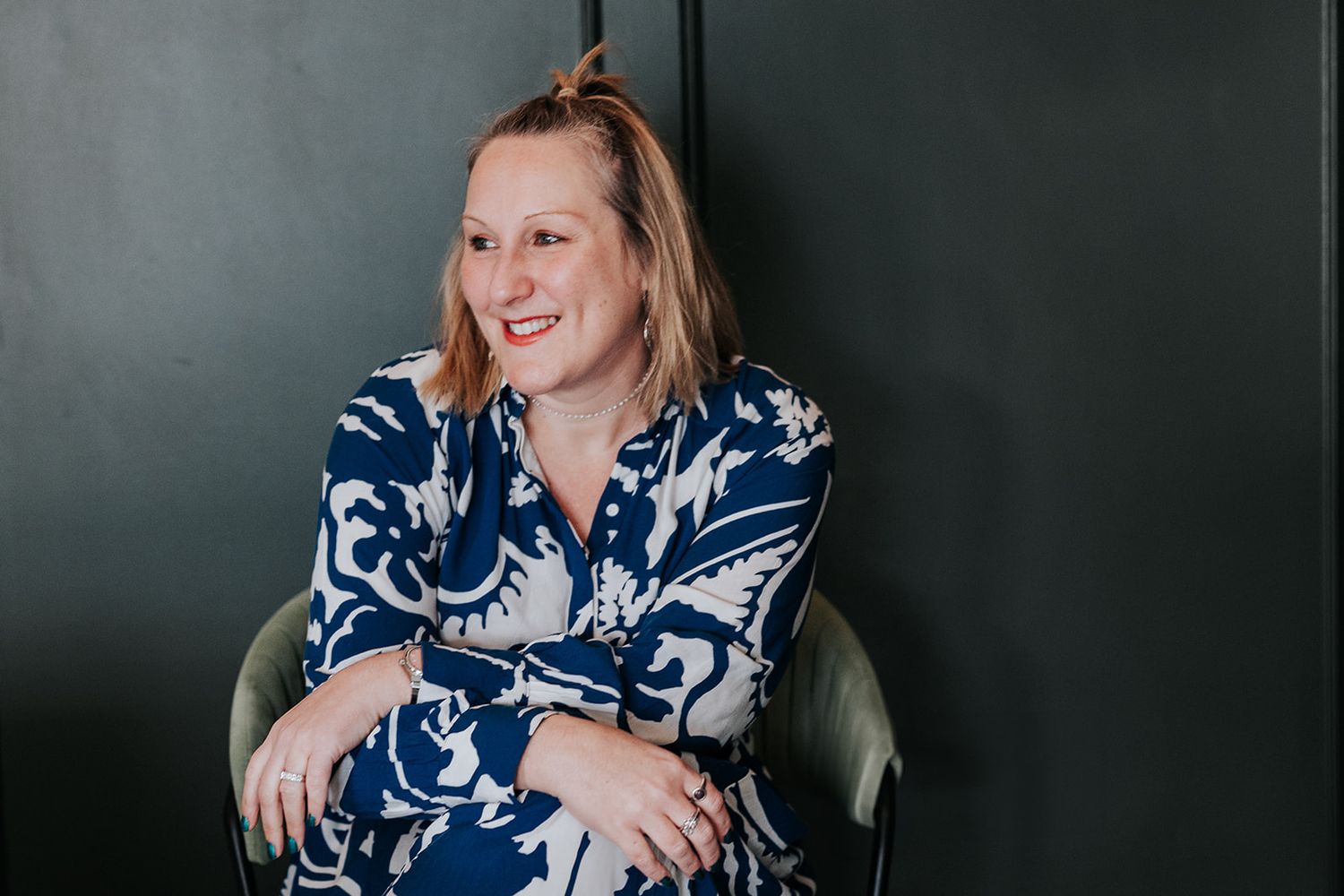 A smiling woman in a navy blue and white patterned dress sits against a dark wall in a styled portrait setting.