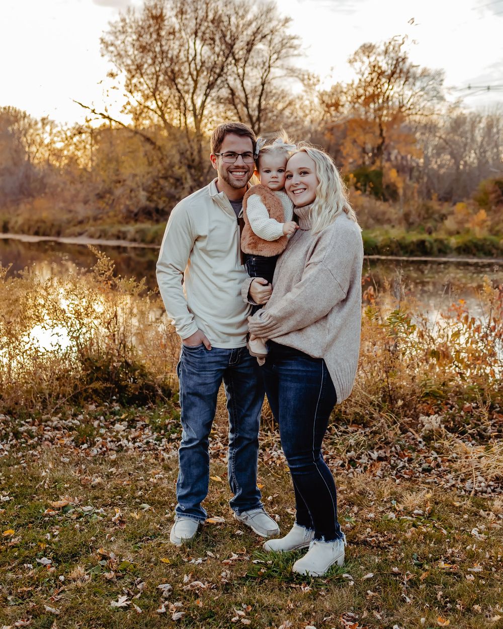 Family in cozy sweaters posing together at sunset by a lake in autumn with golden trees in the background.