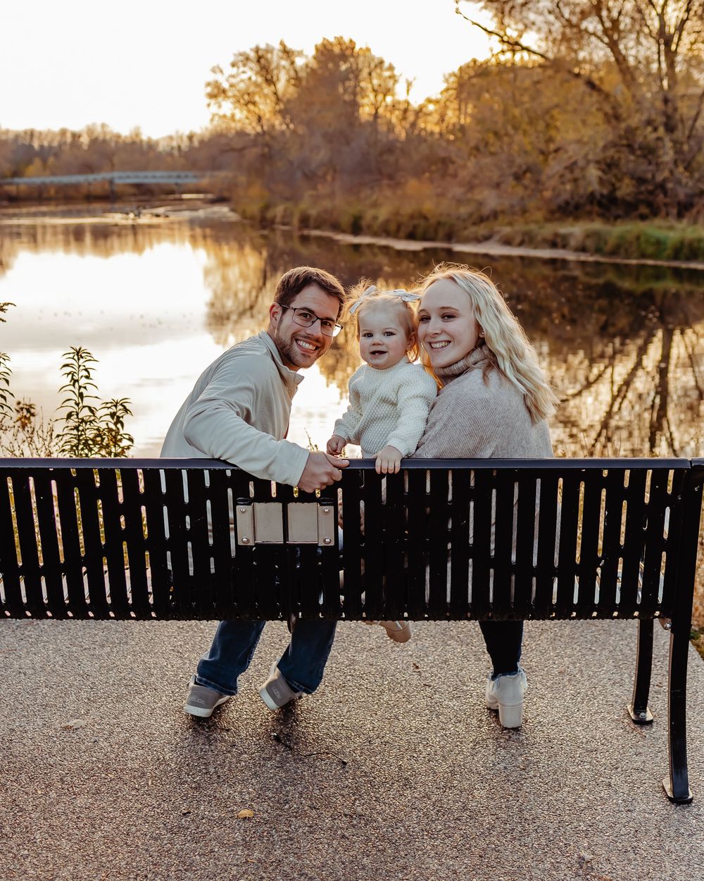 A family sits together on a park bench at sunset by a tranquil lake with autumn trees reflecting in the water.