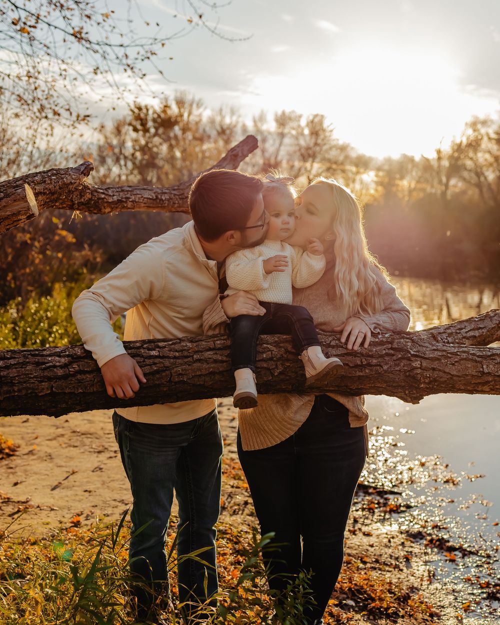 A romantic couple shares a tender moment leaning over a fallen log by a lake at sunset with autumn foliage in the background.