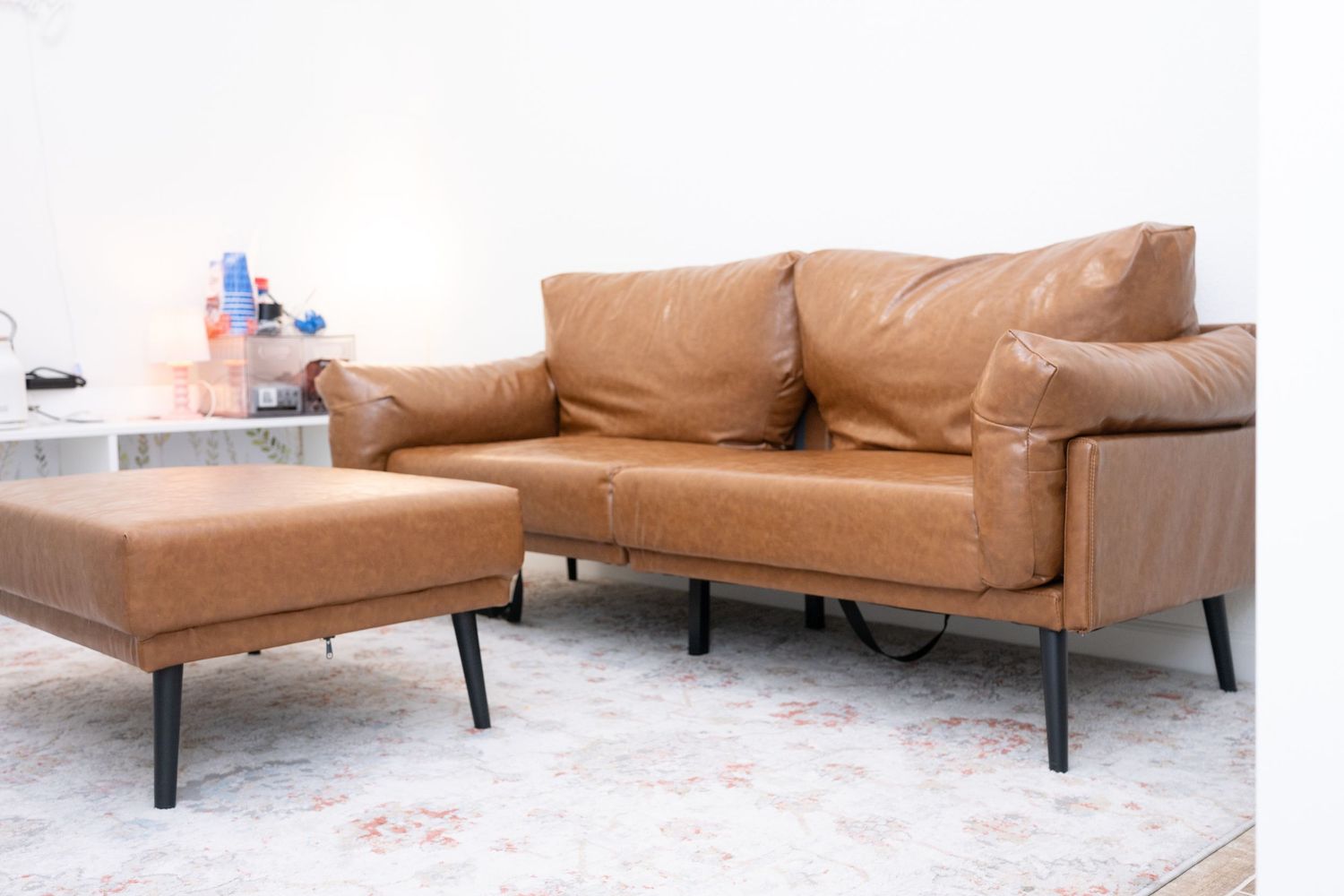 Modern tan leather sofa and ottoman set with black metal legs on concrete floor.