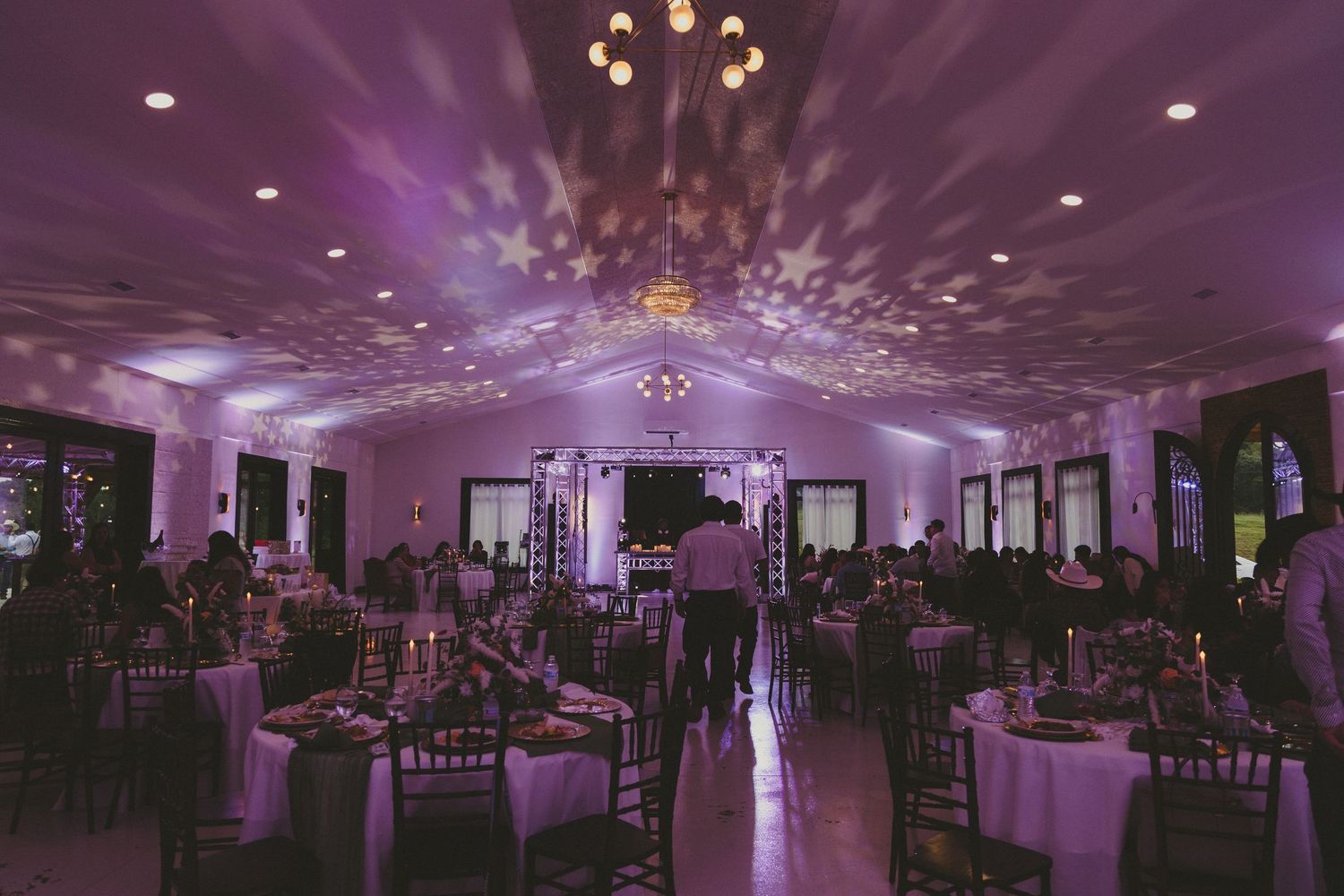Elegant ballroom with purple uplighting features round dining tables, chandeliers and dramatic ceiling lighting patterns.
