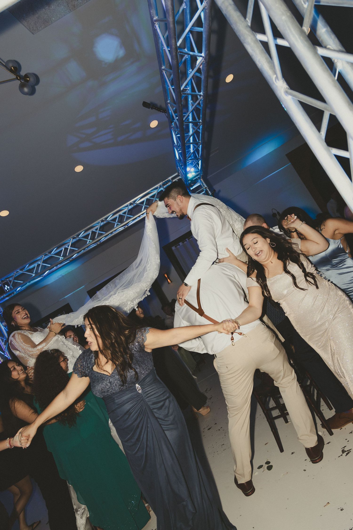 Dancers in formal attire move energetically on a dance floor under blue lighting and overhead truss structures.