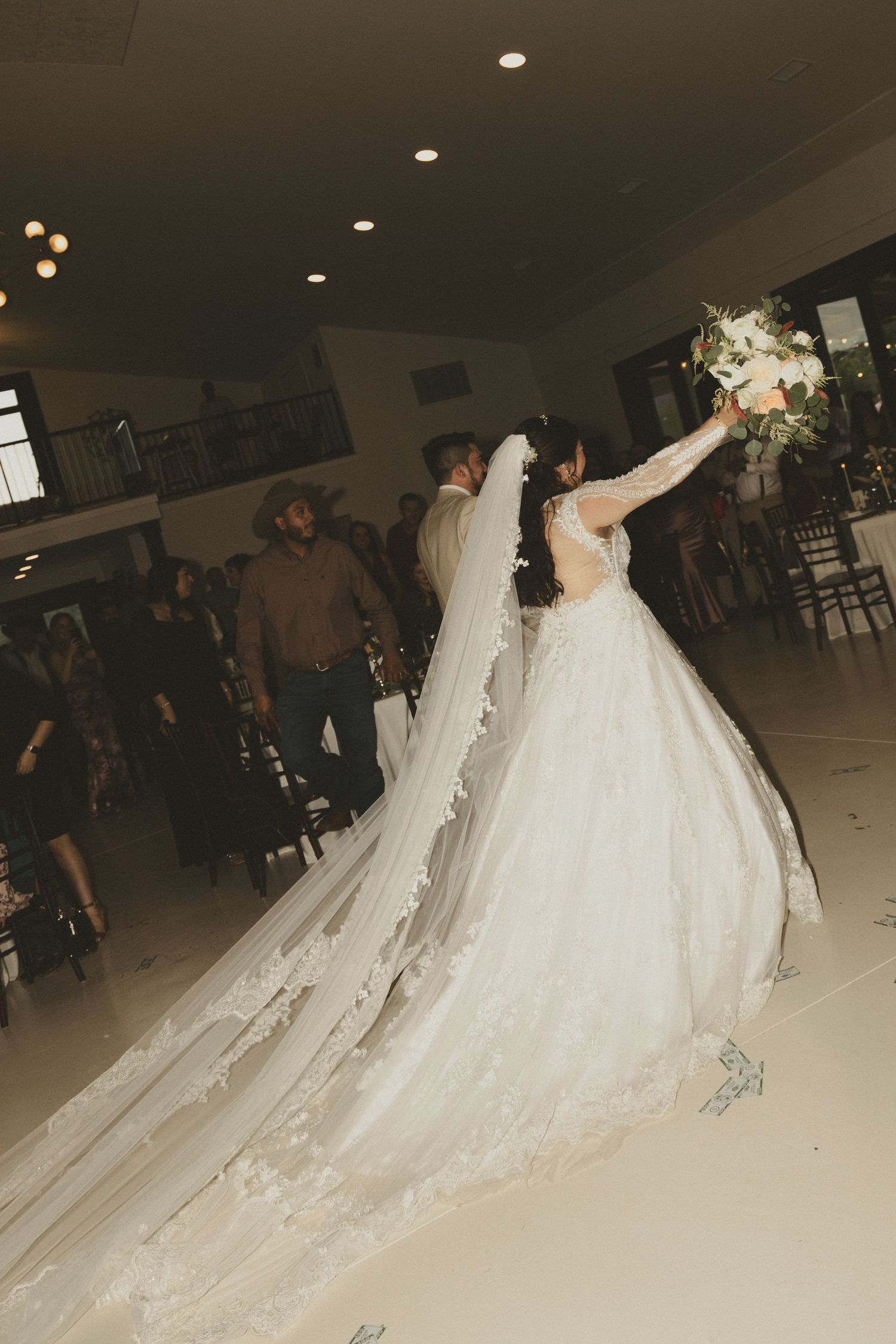 A bride in a flowing white ballgown with a dramatic cathedral-length veil raises her bouquet while dancing at a reception.