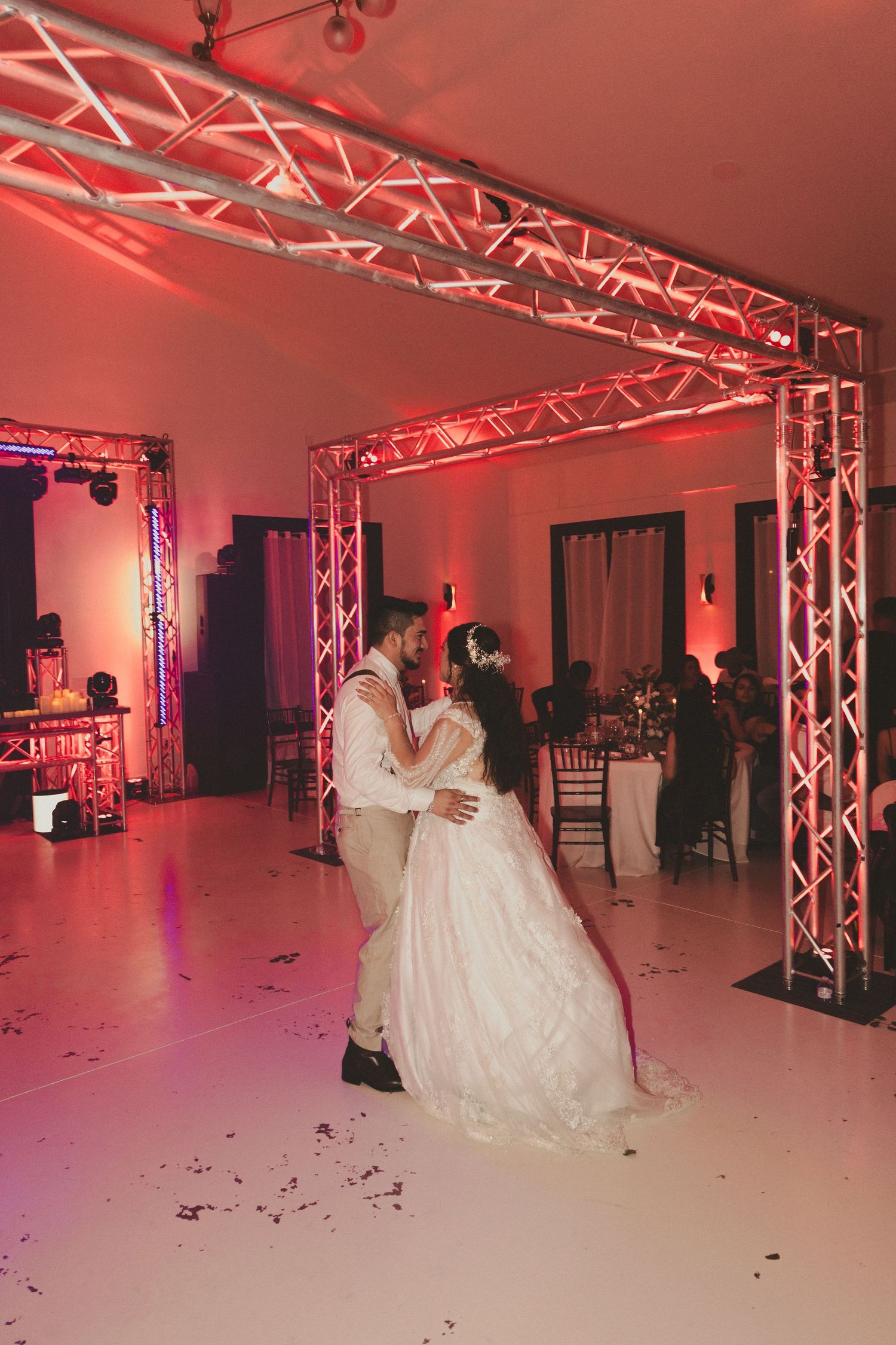 Newlyweds share their first dance in an elegant venue with pink lighting and structural truss beams overhead.