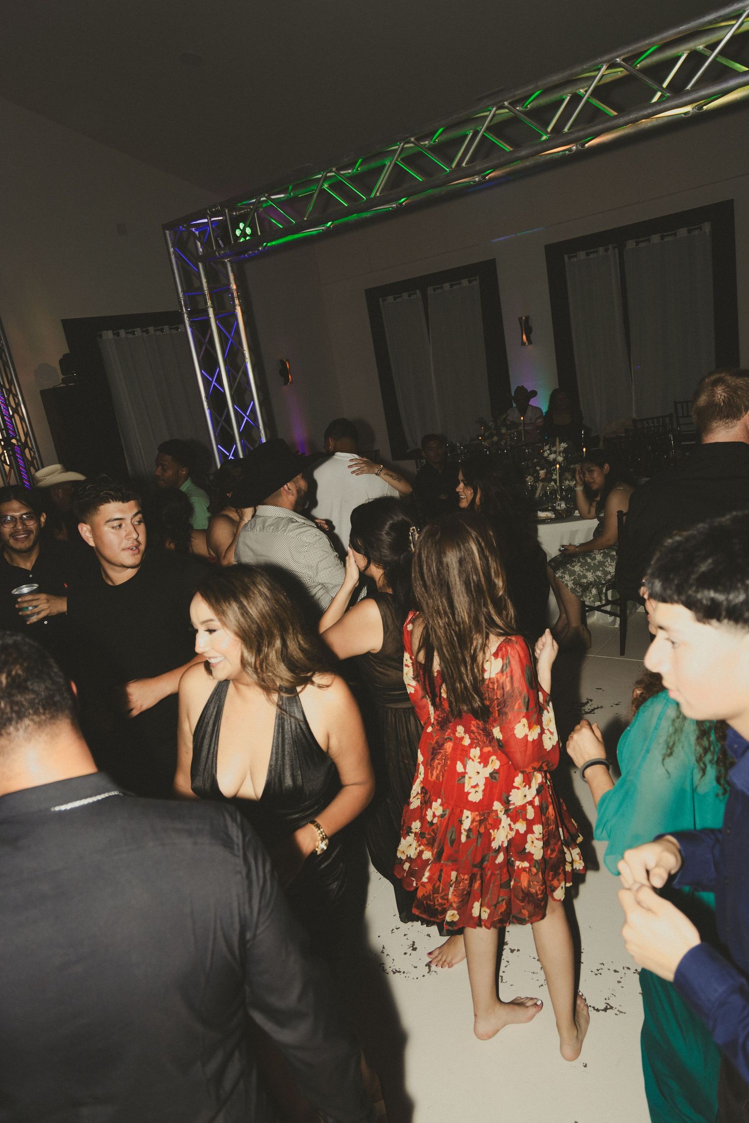 A crowded dance party scene with people dancing under colored stage lighting and truss rigging.