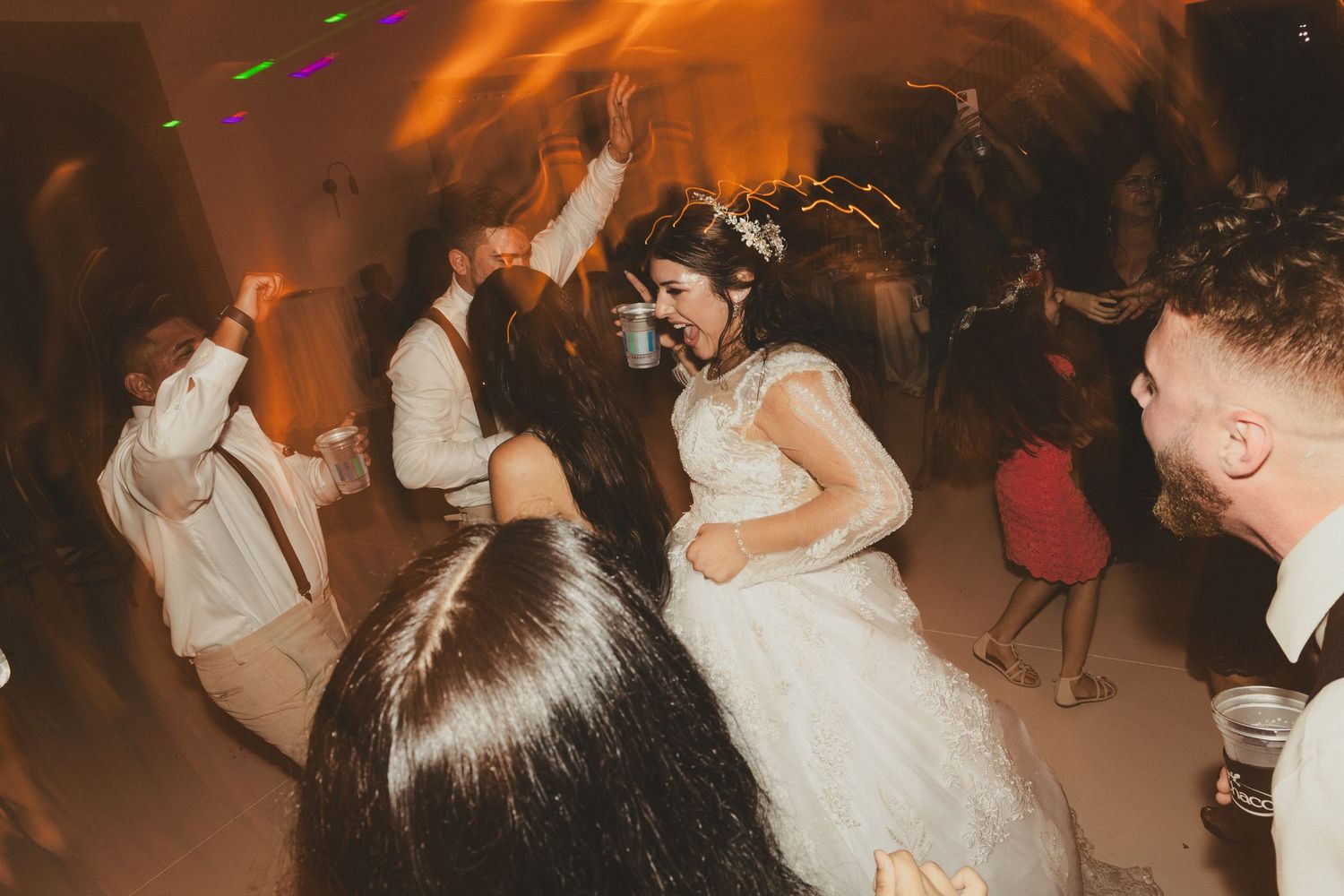 People dancing and celebrating at a wedding reception with warm orange lighting creating a lively party atmosphere.