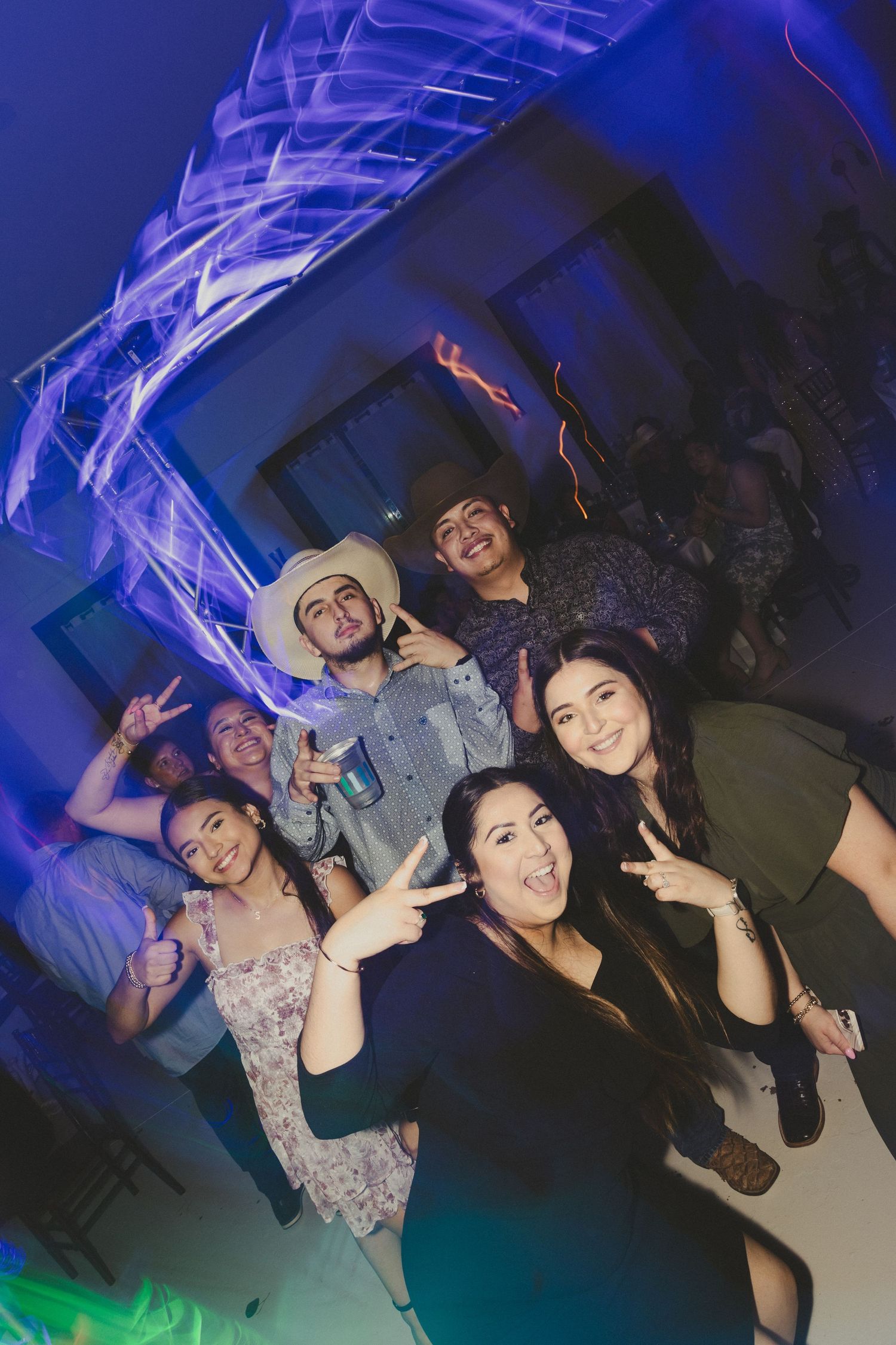 Group of friends posing and making peace signs at a party with blue and green lighting effects in the background.
