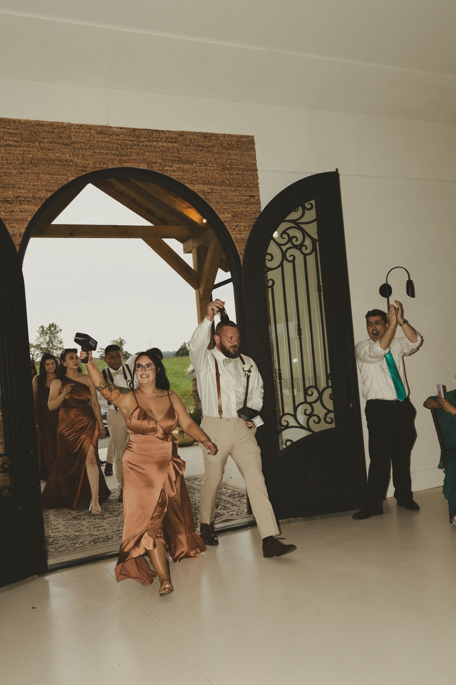 Wedding party members walk through an elegant arched doorway in formal attire with bridesmaids in blush gowns.