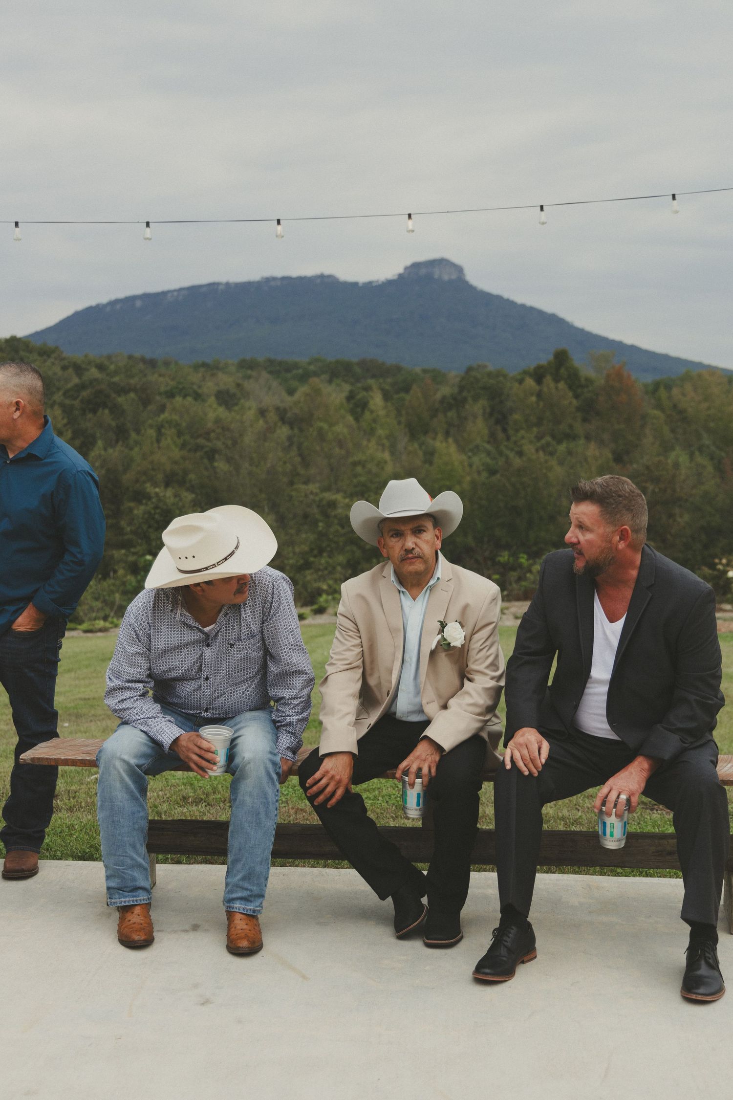 A group of cowboys sit casually on a fence with mountains visible in the background against an overcast sky.