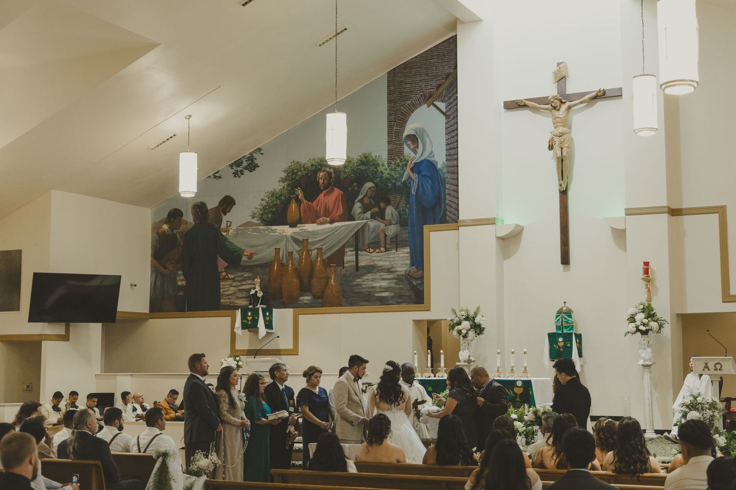 Wedding ceremony taking place in a Catholic church with guests seated beneath religious artwork and a crucifix.