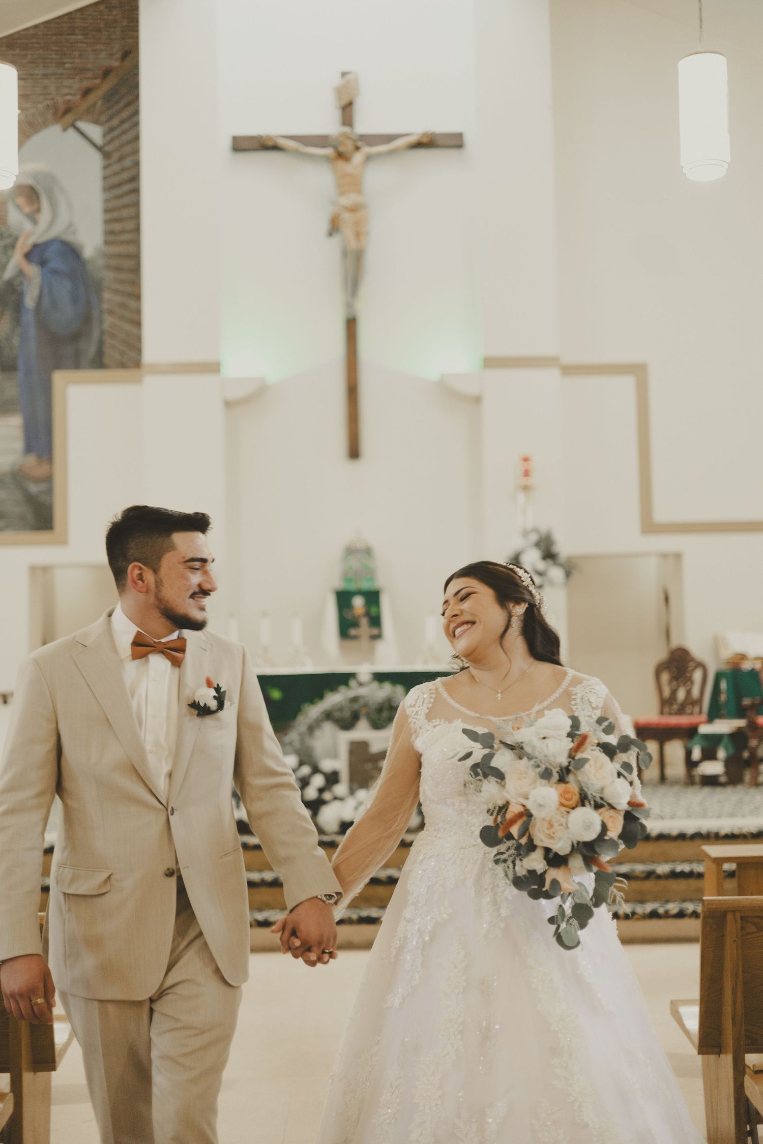 Happy bride in white lace gown and groom in beige suit holding hands and laughing during church ceremony.